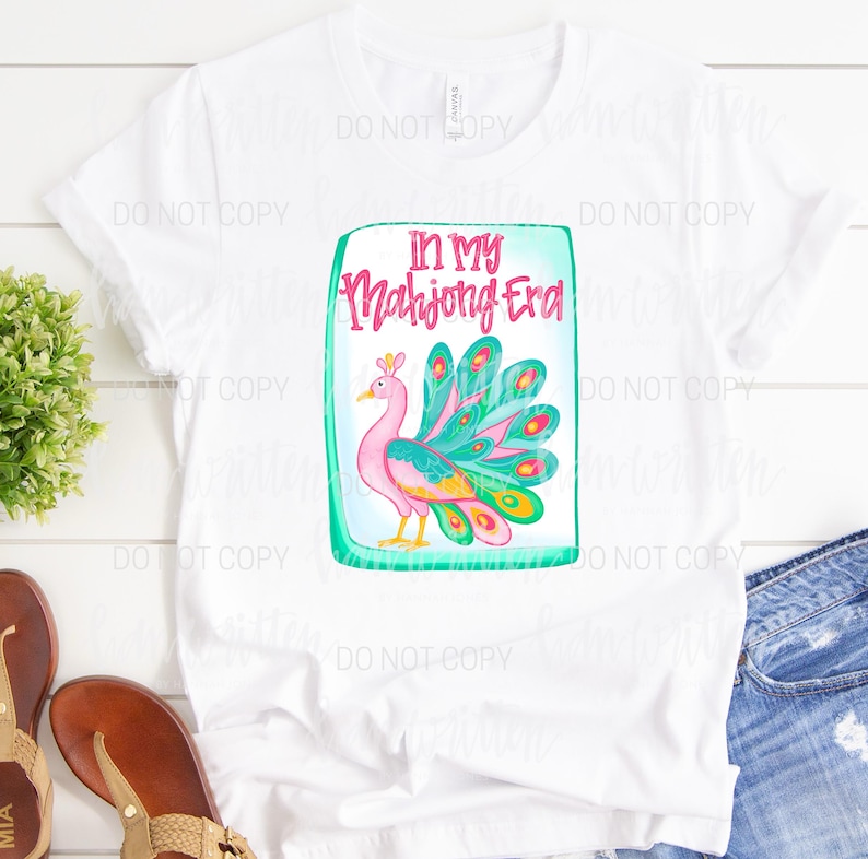 May include: White t-shirt featuring a colorful peacock design inside a green rectangle. The text "IN MY Mahjong Era" is above the peacock. The peacock has pink, teal, yellow, and blue feathers.