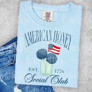May include: Light blue t-shirt with the words "AMERICAN HONEY SOCIAL CLUB" in navy blue script. The shirt features a graphic of a jar with blue flowers and a small American flag. The shirt is paired with blue denim.