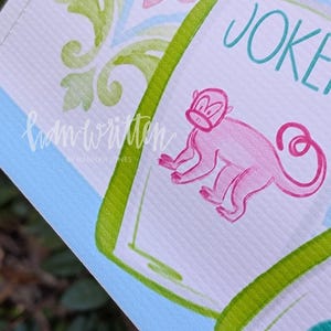 May include: Close-up of a card with a pink monkey illustration and the word "JOKER" in teal. The card has a green border and is set against a backdrop of blue, pink, and green floral patterns. The text "handwritten by Hannah Jones" is also visible.