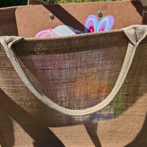 May include: A large brown hessian tote bag with a light brown handle. Inside the bag is a brown clipboard with colourful bunny-shaped decorations. The bag is sitting on green grass.
