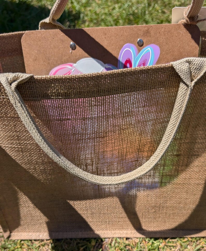 May include: A large brown hessian tote bag with a light brown handle. Inside the bag is a brown clipboard with colourful bunny-shaped decorations. The bag is sitting on green grass.