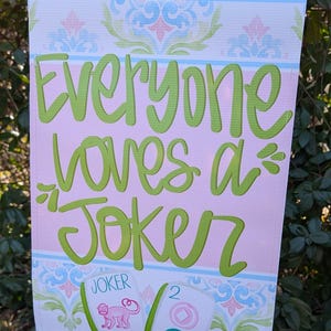 May include: A decorative garden flag with the text "Everyone loves a Joker" in green script on a pink background. The flag has a blue border and features a floral design. Two Mahjong tiles are shown.