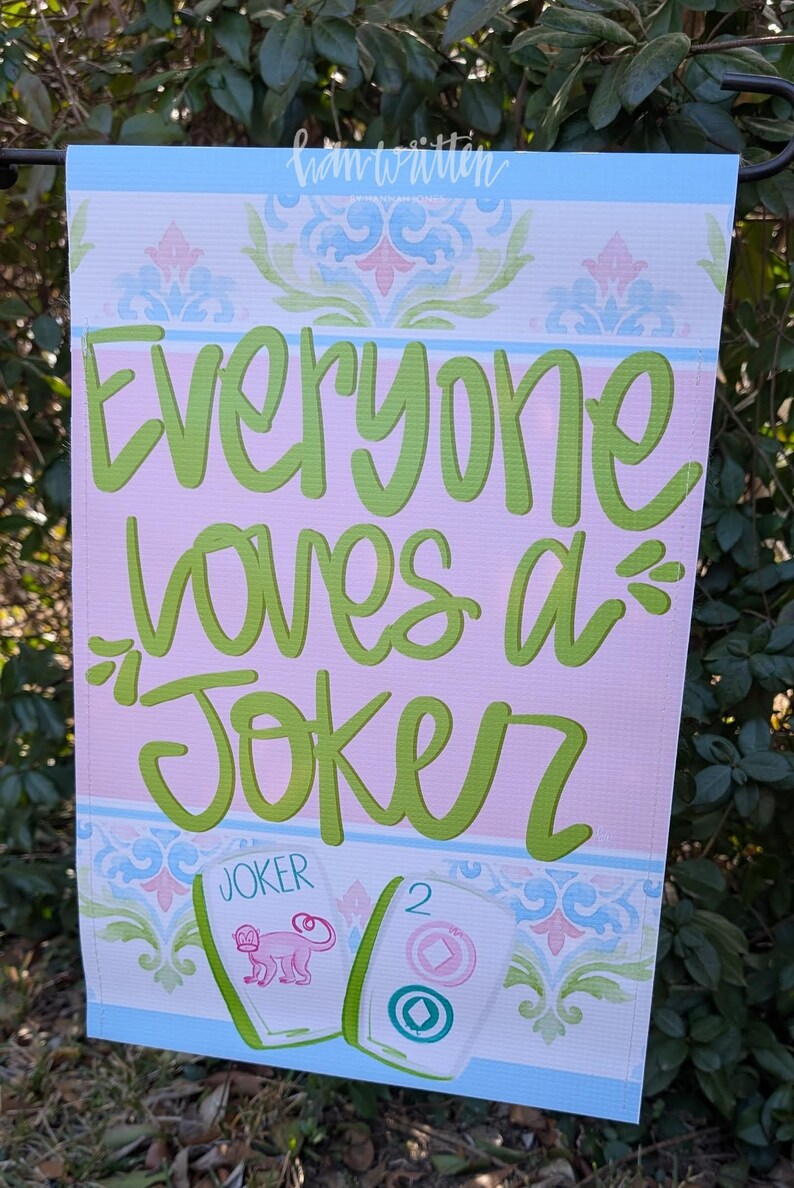 May include: A decorative garden flag with the text "Everyone loves a Joker" in green script on a pink background. The flag has a blue border and features a floral design. Two Mahjong tiles are shown.