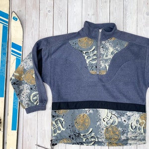 May include: A blue, grey, and black fleece pullover with a half-zip closure. The fleece has a geometric pattern with gold accents on the sleeves and bottom.