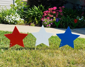 May include: Three wooden stars in red, white, and blue, standing in a row on a green lawn. The stars are a patriotic decoration for the 4th of July.