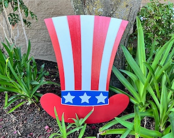 May include: A red, white, and blue wooden Uncle Sam hat decoration. The hat is in the shape of a top hat with a white band and five white stars on the band. The hat is sitting on a bed of brown mulch.