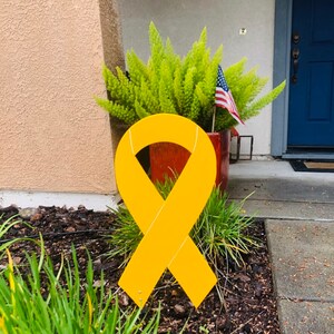 May include: A large yellow ribbon made of metal, standing in a yard with green plants and brown mulch. The ribbon is a symbol of support and awareness.