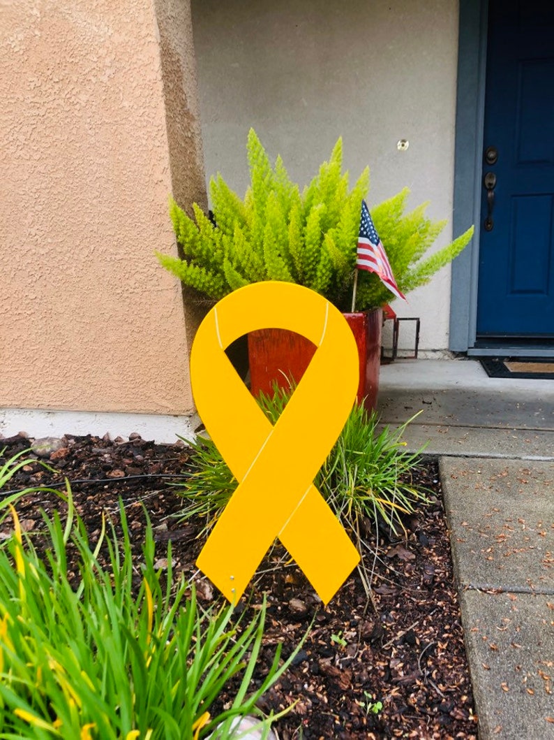 May include: A large yellow ribbon made of metal, standing in a yard with green plants and brown mulch. The ribbon is a symbol of support and awareness.
