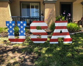May include: Large wooden letters spelling "USA" painted in red, white, and blue stripes, resembling the American flag. The letters are displayed on a grassy lawn.