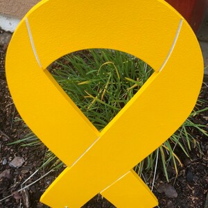 May include: A yellow ribbon made of wood with white lines. The ribbon is in the shape of a bow and is standing upright in the ground.