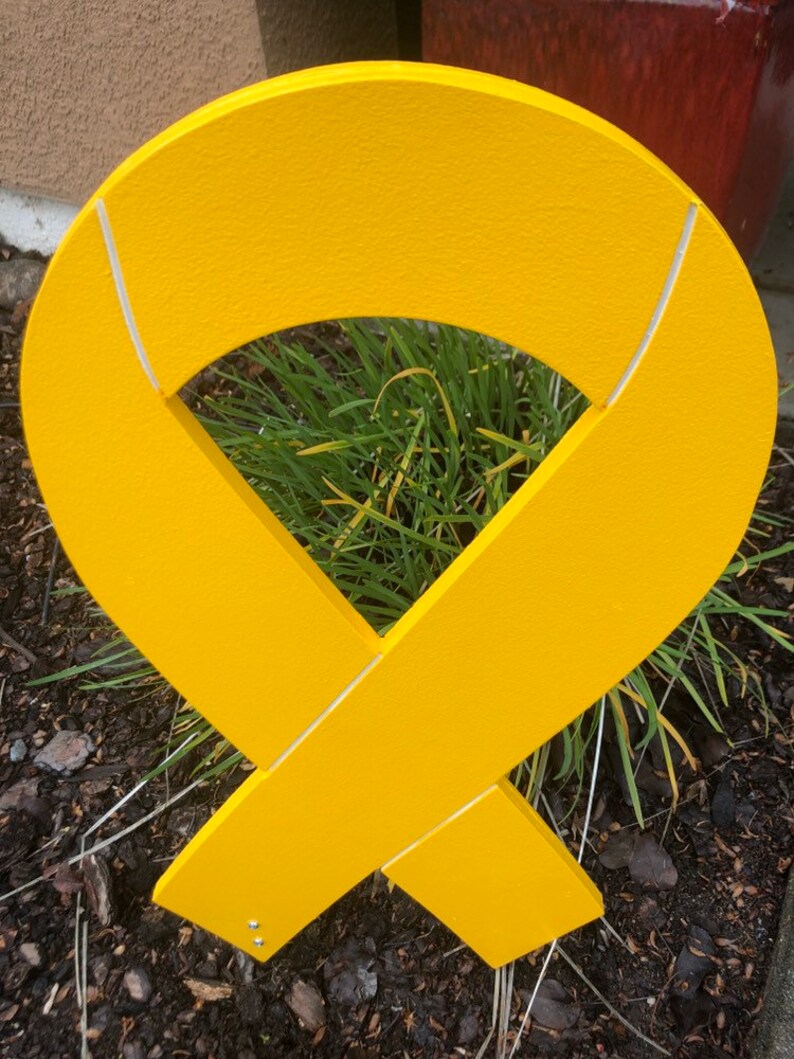 May include: A yellow ribbon made of wood with white lines. The ribbon is in the shape of a bow and is standing upright in the ground.