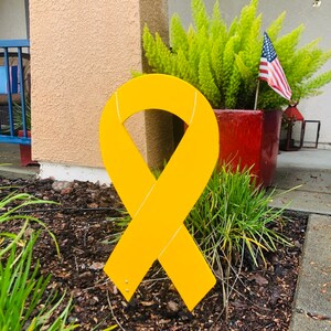 May include: A large yellow ribbon made of wood, standing upright in a bed of brown mulch. The ribbon is tied in a bow and is a symbol of support and awareness.