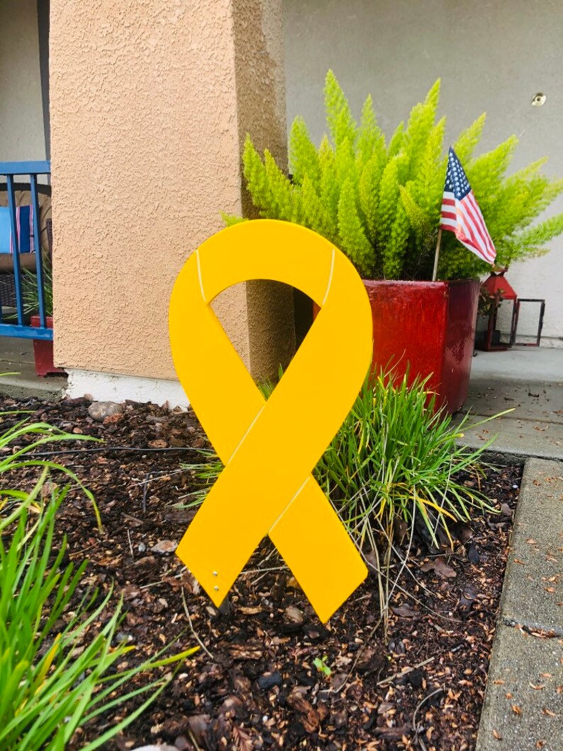 May include: A large yellow ribbon made of wood, standing upright in a bed of brown mulch. The ribbon is tied in a bow and is a symbol of support and awareness.