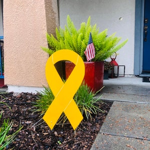 May include: A large yellow ribbon is displayed on a lawn in front of a house. The ribbon is made of a sturdy material and is designed to be displayed outdoors.
