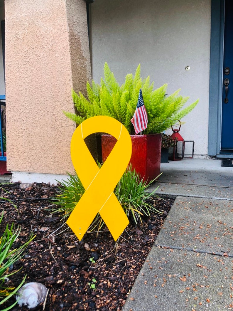 May include: A large yellow ribbon is displayed on a lawn in front of a house. The ribbon is made of a sturdy material and is designed to be displayed outdoors.