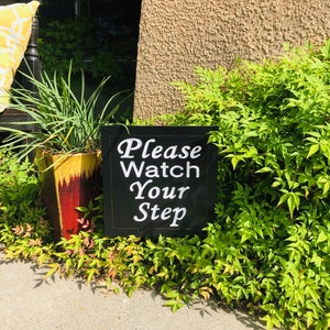 May include: A black sign with white text that reads "Please Watch Your Step" is placed in front of a bush. The sign is in front of a brown stucco wall.