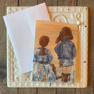 May include: A greeting card featuring a watercolor illustration of two girls wearing denim jackets and jeans. The girls are standing back to back with their backs to the viewer. The card has a light orange background and the artist's signature in the bottom right corner.