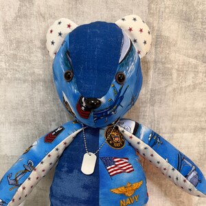 May include: A stuffed teddy bear constructed from various blue fabric patches featuring United States Navy themes, including aircraft, anchors, the American flag, and the word NAVY. The bear has white ears and feet with red and blue star patterns and wears a silver-toned ball chain necklace with a blank rectangular metal dog tag.