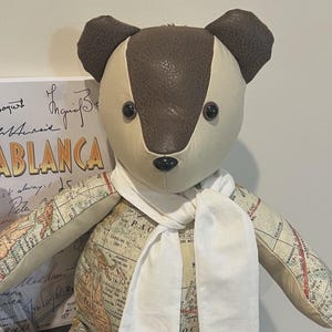May include: A stuffed bear toy constructed from a map-patterned fabric on the body and limbs, with solid cream and dark brown textured panels on the head and ears. The bear features black button eyes, a black nose, and a plain white fabric scarf.