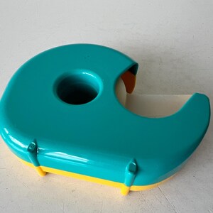 May include: A teal and yellow tape dispenser. The dispenser has a rounded, ergonomic design with a large central hole and a curved cutting edge. The base is yellow, and the tape roll is visible.