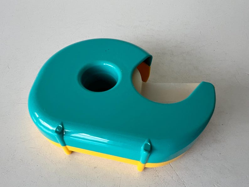 May include: A teal and yellow tape dispenser. The dispenser has a rounded, ergonomic design with a large central hole and a curved cutting edge. The base is yellow, and the tape roll is visible.