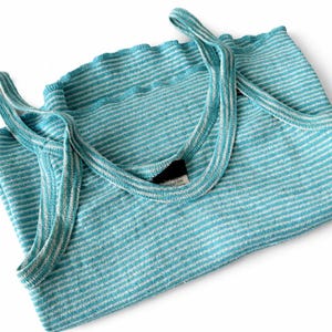 Puede incluir: Una camiseta sin mangas a rayas turquesa y blancas doblada con tirantes finos. La parte superior est&aacute; hecha de una tela suave y texturizada y tiene un escote festoneado. Los tirantes son de color turquesa a juego. La camiseta sin mangas est&aacute; doblada cuidadosamente sobre un fondo blanco.