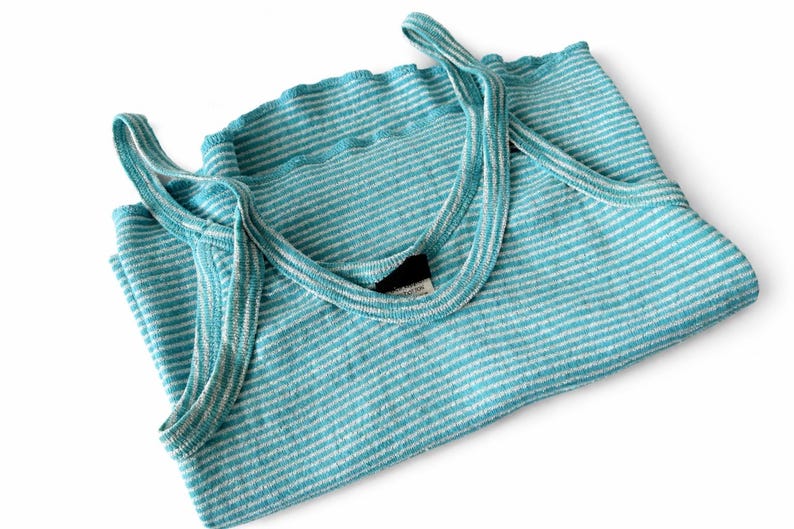 Puede incluir: Una camiseta sin mangas a rayas turquesa y blancas doblada con tirantes finos. La parte superior est&aacute; hecha de una tela suave y texturizada y tiene un escote festoneado. Los tirantes son de color turquesa a juego. La camiseta sin mangas est&aacute; doblada cuidadosamente sobre un fondo blanco.