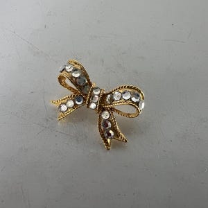 May include: A gold-toned bow brooch adorned with clear, sparkling rhinestones. The bow design features intricate detailing and a textured appearance, creating a glamorous accessory. The brooch is set against a neutral background.