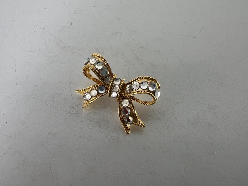 May include: A gold-toned bow brooch adorned with clear, sparkling rhinestones. The bow design features intricate detailing and a textured appearance, creating a glamorous accessory. The brooch is set against a neutral background.