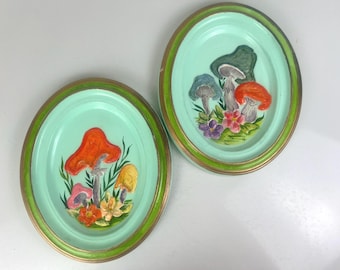 May include: Two decorative plates with a green rim and a light blue center. Each plate features a 3D design of mushrooms and flowers in shades of red, orange, yellow, green, and purple.