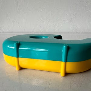 May include: A teal and yellow tape dispenser. The dispenser is a rounded rectangular shape with a teal top and yellow base. It has a circular opening on top for the tape roll.