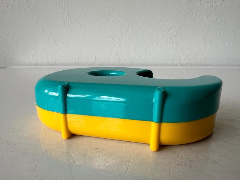 May include: A teal and yellow tape dispenser. The dispenser is a rounded rectangular shape with a teal top and yellow base. It has a circular opening on top for the tape roll.
