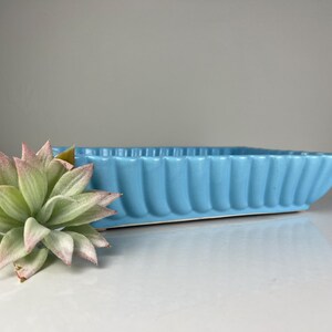 May include: A light blue ceramic planter with a fluted design. The planter is rectangular and has a smooth, glossy finish.