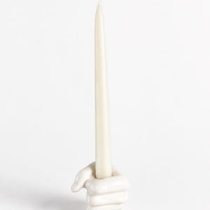 May include: A white ceramic candle holder shaped like a hand, holding a long, off-white taper candle. The candle is slender and has a slightly curved wick. The holder is smooth and detailed, with visible fingers and a thumb.