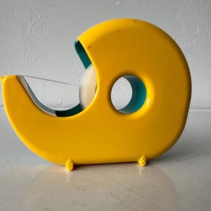 May include: A bright yellow tape dispenser with a teal interior. The dispenser has a rounded shape with a circular opening and a blade for cutting tape. The tape roll is visible inside the dispenser.