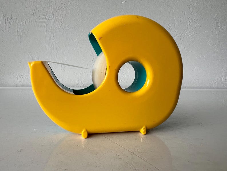 May include: A bright yellow tape dispenser with a teal interior. The dispenser has a rounded shape with a circular opening and a blade for cutting tape. The tape roll is visible inside the dispenser.