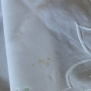 May include: White linen tablecloth with embroidered floral design in shades of blue, green, yellow, and pink. There is a small brown stain on the tablecloth.