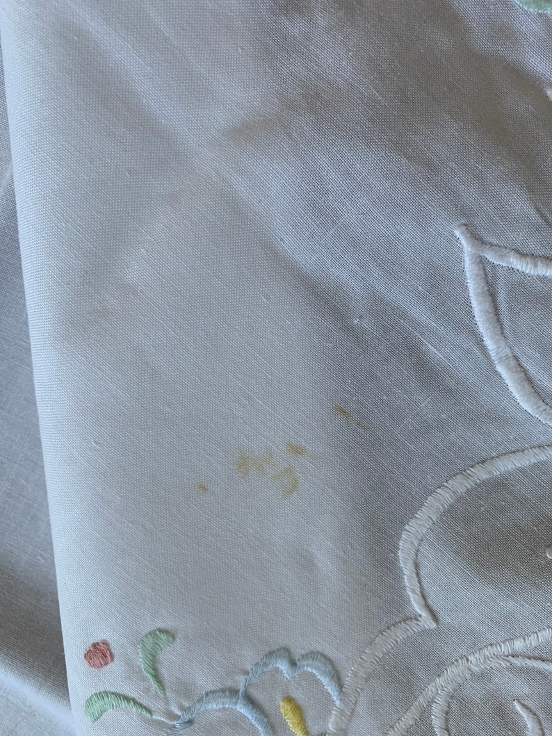 May include: White linen tablecloth with embroidered floral design in shades of blue, green, yellow, and pink. There is a small brown stain on the tablecloth.