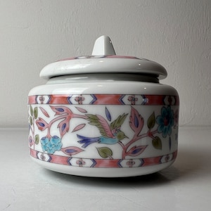 May include: A white ceramic jar with a lid, featuring a floral design with pink, blue, and green accents. The jar has a stylized bird design on the front.