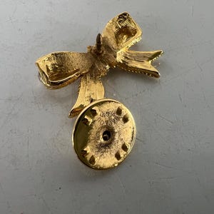 May include: A gold-colored bow pin with a textured surface. The pin has a bow shape with detailed folds and a round backing with a pin closure. The pin is likely made of metal and is designed to be attached to clothing or accessories.