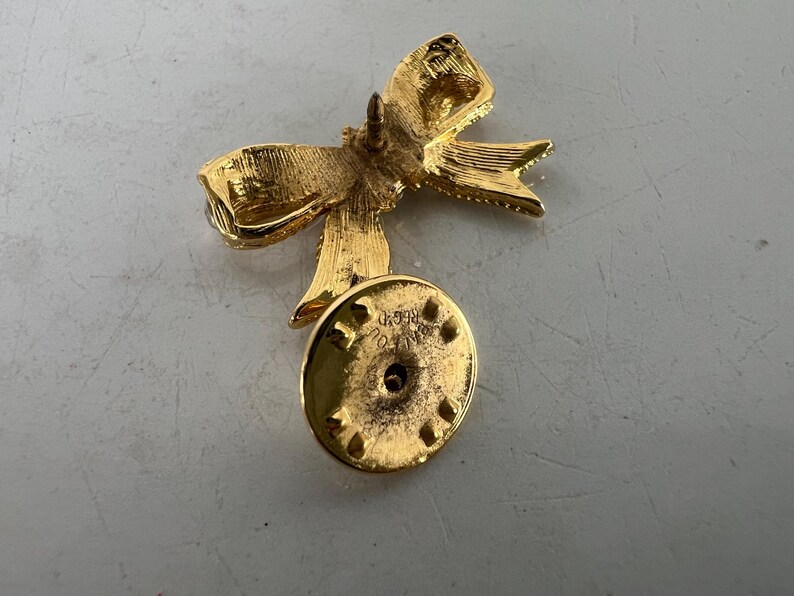 May include: A gold-colored bow pin with a textured surface. The pin has a bow shape with detailed folds and a round backing with a pin closure. The pin is likely made of metal and is designed to be attached to clothing or accessories.