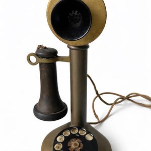 May include: Antique brass rotary dial telephone with a black earpiece and mouthpiece. The phone has a circular base and a vertical stand supporting the earpiece. The dial features letters and numbers. A cord extends from the base.