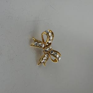 May include: A gold-toned bow brooch adorned with clear, sparkling rhinestones. The bow design features intricate detailing and a classic shape, perfect for adding a touch of elegance to any outfit. This jewelry piece is a timeless accessory.