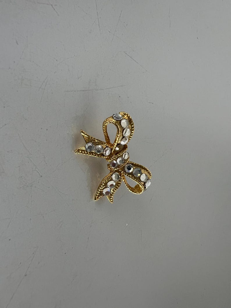 May include: A gold-toned bow brooch adorned with clear, sparkling rhinestones. The bow design features intricate detailing and a classic shape, perfect for adding a touch of elegance to any outfit. This jewelry piece is a timeless accessory.