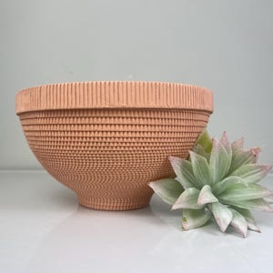 May include: A light pink ceramic planter with a textured, geometric pattern. The planter is empty and has a wide, shallow bowl shape.