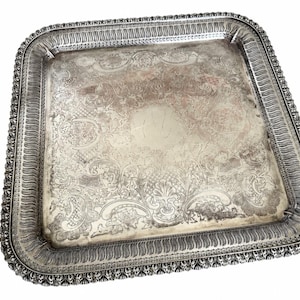 May include: A square silver-toned serving tray featuring an ornate, embossed floral and scrollwork pattern across the surface. The tray has a raised, decorative border with a repeating scalloped edge and intricate detailing along the rim.