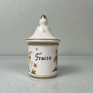 May include: White ceramic jar with a green lid and a green and yellow floral design. The jar is labeled "Fraise" in black lettering.