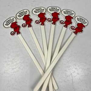 May include: Six white plastic cocktail stirrers with red cartoon devil figures on the top. Each devil figure has a speech bubble that says "Here's something tempting!"