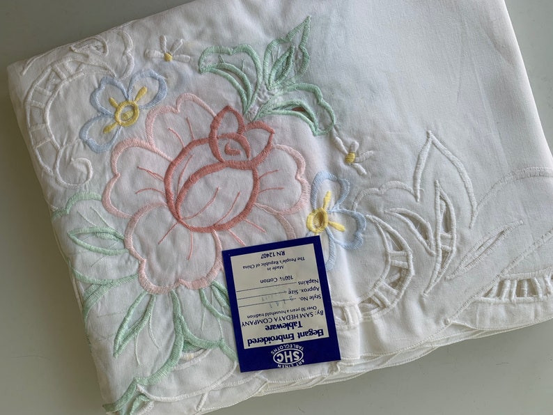 May include: A white embroidered tablecloth featuring a floral design with pink, blue and yellow flowers. The tablecloth is made from 100% cotton and has a label that reads "Begant Embroidered Tableware by Sam Hedaya Company, Approx Size Napkins, 100% Cotton, RN 12407".