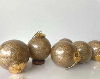 May include: Several gold glitter Christmas ornaments with gold-colored metal caps and hangers. One ornament is pear-shaped and sits on a small wooden base. The other ornaments are round and vary in size. The ornaments are on a white surface.